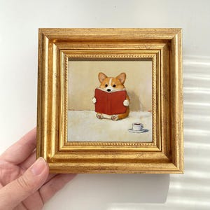 May include: A framed painting depicts a corgi dog reading a red book, with a small cup of coffee nearby. The painting is set in a gold frame. The corgi is brown and white, and the book is a vibrant red. The painting has a warm, inviting feel.