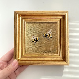 May include: A square painting featuring two bumblebees with yellow and black striped bodies and translucent wings, set against a textured gold background. The artwork is encased in an ornate, metallic gold frame with a stepped, beveled edge design.
