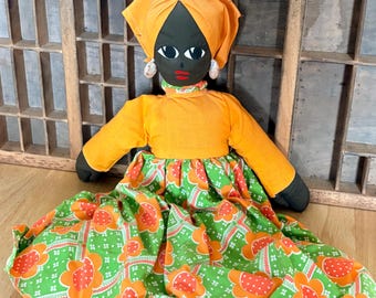 May include: A handmade doll with a black face, wearing an orange head wrap and top, and a green skirt with orange floral patterns. The doll has white earrings and a red mouth. The doll's arms and legs are black.