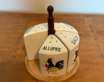May include: A vintage spice rack with four white ceramic containers labeled "Salt", "Allspice", "Cinnamon", and "Pepper". Each container has a hole-punched top and a painted rooster design. The rack sits on a wooden base and has a wooden handle.