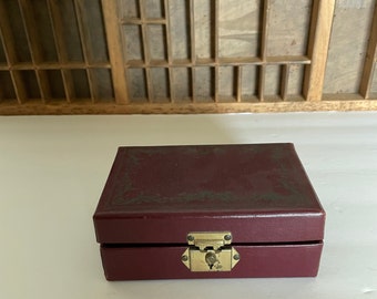 May include: A burgundy leather-bound box with a gold latch and decorative details. The box is closed and sitting on a white surface.