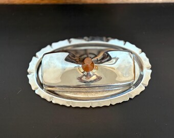 May include: A silver-colored butter dish with a rectangular lid and an oval base. The lid has a small, round, amber-colored knob. The base has a scalloped edge.