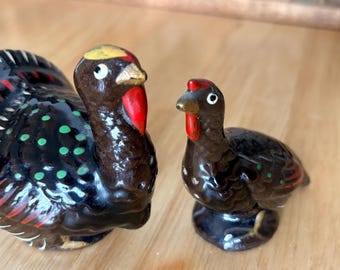 May include: A pair of ceramic turkey figurines. The larger turkey is dark brown with a black tail, green spots, and red accents. The smaller turkey has similar coloring. Both have red wattles and yellow beaks. They are likely salt and pepper shakers.