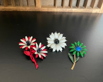 May include: Three enamel flower brooches. One white flower with a black center, one red and white flower with a black center, and one green flower with a blue center.