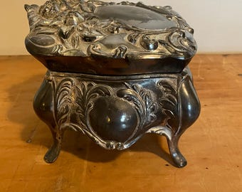 May include: A silver-plated trinket box with ornate floral and scrollwork designs. The box has a hinged lid and sits on four decorative feet.