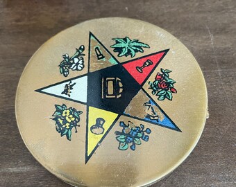 May include: A gold-colored compact with a five-pointed star design. The star is divided into five sections, each with a different symbol and color. The symbols include a trowel, a plumb bob, a level, a book, and a compass. The compact is likely a Masonic symbol.