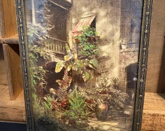 May include: A framed vintage print depicting a European courtyard scene with lush greenery, a balcony, and a red awning. The frame is ornate, with a dark, metallic finish. The artwork evokes a sense of nostalgia and classic decor.