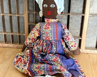May include: A handmade doll with a dark complexion, wearing a white head wrap, and a floral print dress with purple accents. The doll has red lips and is seated on a wooden surface. Light blue shoes are placed in front of the doll.