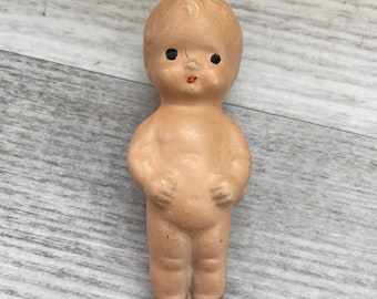 May include: A vintage porcelain doll with a painted face and black eyes. The doll is nude and has a simple, childlike appearance.