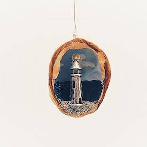Pode incluir: Uma miniatura de farol em tom prateado colocada dentro de uma casca de noz oca. O interior apresenta um fundo pintado com mar azul escuro e céu azul claro, com uma base de areia texturizada na parte inferior da casca.
