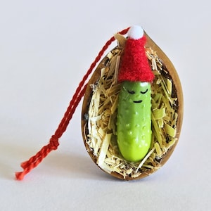 May include: A quirky Christmas decoration. A green pickle with a red Santa hat rests in a wooden shell filled with straw. The ornament has a red string for hanging.