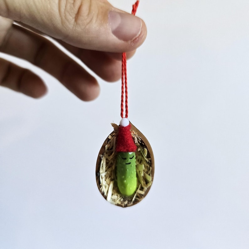 May include: A quirky Christmas bauble showing a green pickle wearing a red Santa hat. The pickle is nestled in a gold-coloured shell with straw, hung by a red string. A unique festive decoration.
