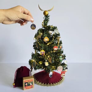May include: A miniature Christmas tree decorated with character ornaments and fairy lights. A golden snitch tops the tree, and a hand holds a small ornament. A red tree skirt and a small box of "Lumos Lights" are at the base.