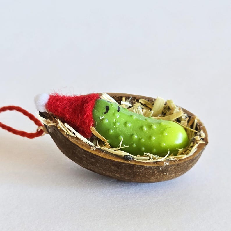 May include: A green pickle Christmas ornament wearing a red Santa hat, resting in a brown shell filled with straw. The pickle has a smiling face. A red string is attached for hanging. Festive holiday decor.