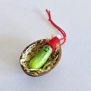 May include: A novelty Christmas tree decoration. A green pickle with a smiling face wears a red Santa hat, resting in a walnut shell filled with straw. A red string is attached for hanging.