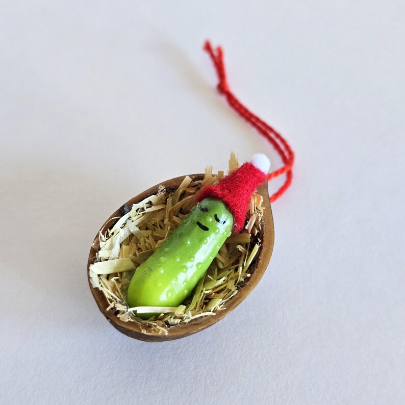 May include: A novelty Christmas tree decoration. A green pickle with a smiling face wears a red Santa hat, resting in a walnut shell filled with straw. A red string is attached for hanging.