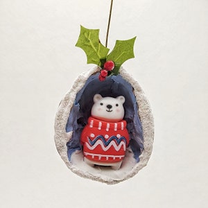 May include: A white polar bear wearing a red and blue sweater sits inside a white, snow-covered, hollowed-out walnut shell ornament. The ornament is topped with a green holly leaf sprig and red berries.