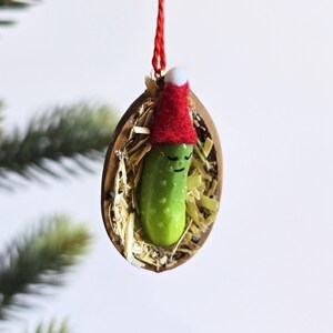 May include: A quirky Christmas decoration. A green pickle with a smiling face, wearing a red Santa hat, nestled in a walnut shell filled with straw, suspended by a red string. A unique festive ornament.