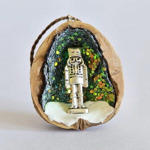 Puede incluir: Adorno navideño artesanal. Una figura de cascanueces plateada se encuentra dentro de una cáscara de nuez, rodeada de purpurina verde y 'nieve' blanca. Tiene una cuerda marrón para colgar. Decoración festiva única.