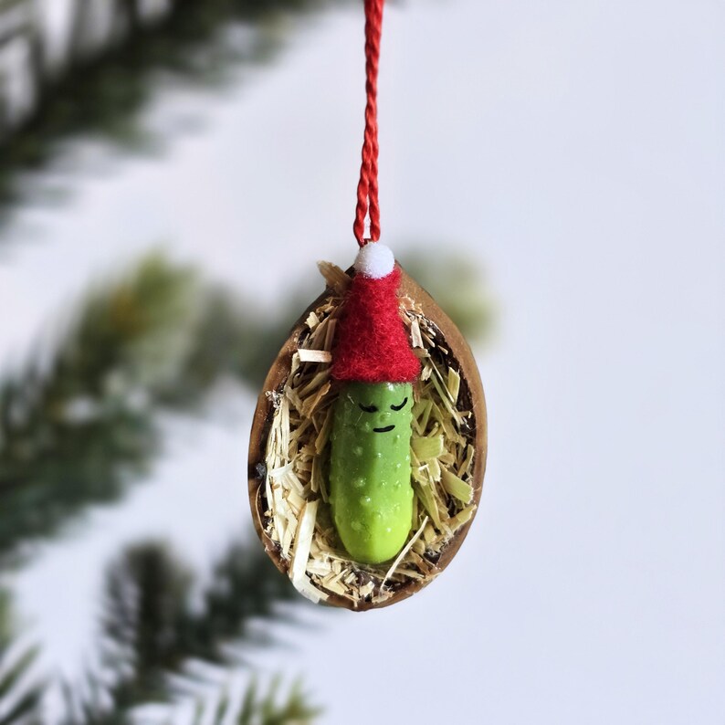 May include: A quirky Christmas decoration. A green pickle wearing a red Santa hat is nestled in a walnut shell filled with straw. The ornament hangs from a red string, ideal for festive decor.