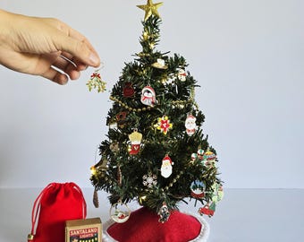 Könnte beinhalten: Ein Miniatur-Weihnachtsbaum, geschmückt mit Ornamenten, einem goldenen Stern und Lichtern. Ein roter Baumrock und ein roter Samtbeutel befinden sich am Fuß. Eine Schachtel mit "Santaland Lights" ist ebenfalls vorhanden. Eine Hand hält einen Mistelzweig-Schmuck.
