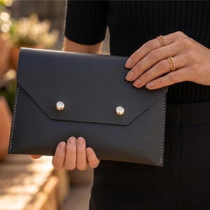May include: Black leather envelope clutch with silver snap closures. The clutch has a textured surface and white stitching along the edges. The bag is held by a person wearing gold jewellery.