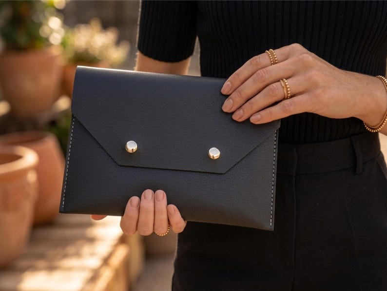 May include: Black leather envelope clutch with silver snap closures. The clutch has a textured surface and white stitching along the edges. The bag is held by a person wearing gold jewellery.