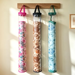 May include: Three cylindrical storage bags hanging from a wooden rack. The left bag is pink with a floral pattern, the centre bag is brown with a large floral print, and the right bag is light blue with a blue floral pattern. Each has a clear top.
