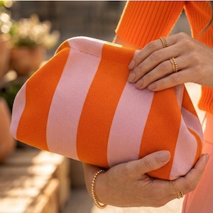 Pode incluir: Uma bolsa clutch estruturada com um padr&atilde;o de malha vibrante com listras verticais alternadas em laranja brilhante e rosa pastel suave. A bolsa tem um design de estrutura arredondada e macia com fecho oculto, exibindo um acabamento de tecido texturizado.