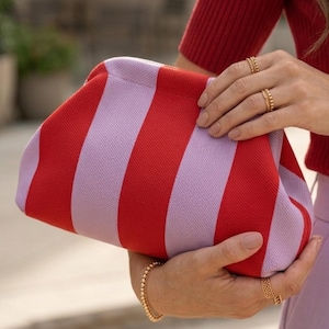 Pode incluir: Uma bolsa clutch com um padr&atilde;o ousado de listras verticais em tons alternados de vermelho vibrante e lavanda suave. A bolsa possui um design de estrutura arredondada com um exterior de tecido texturizado e um fecho superior integrado.