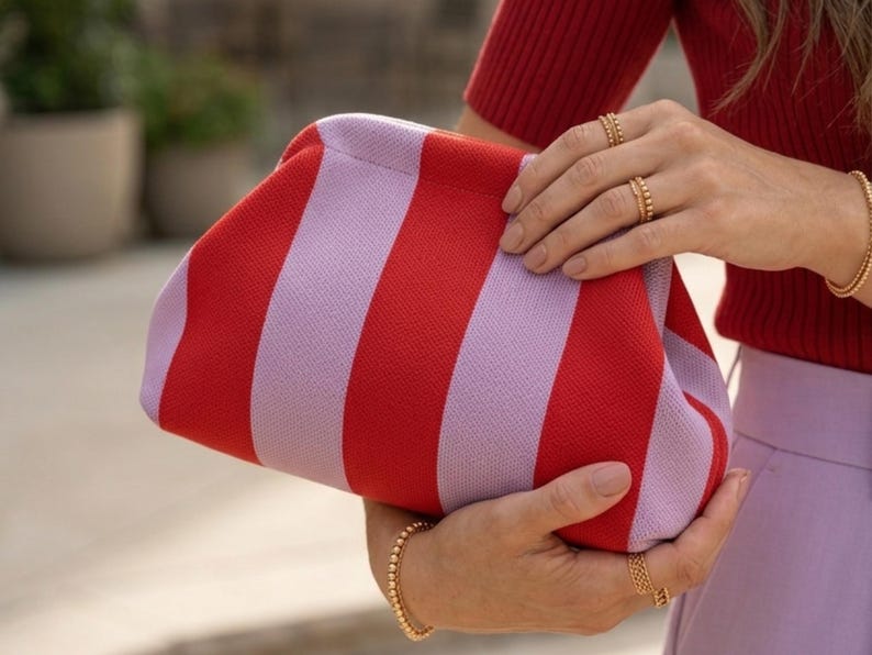 Pode incluir: Uma bolsa clutch com um padr&atilde;o ousado de listras verticais em tons alternados de vermelho vibrante e lavanda suave. A bolsa possui um design de estrutura arredondada com um exterior de tecido texturizado e um fecho superior integrado.