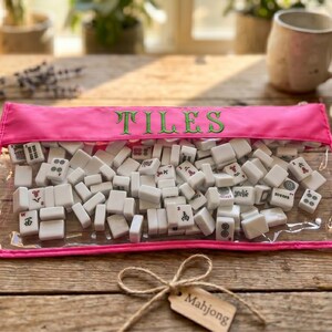 May include: A clear pouch with a pink trim, filled with white Mahjong tiles. The word "TILES" is embroidered in green on the pouch. A small tag with the word "Mahjong" hangs from a twine bow, on a wooden surface.
