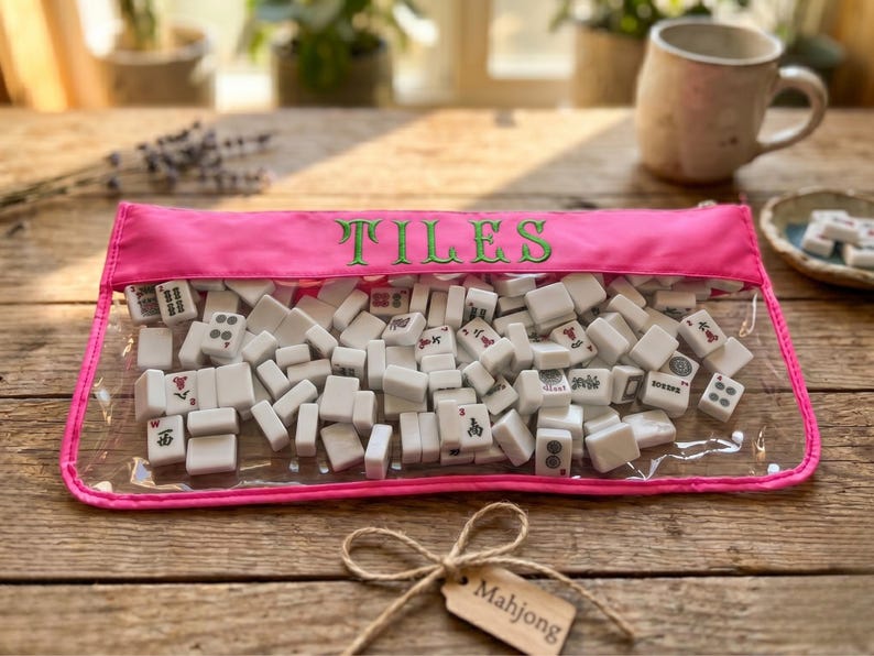 May include: A clear pouch with a pink trim, filled with white Mahjong tiles. The word "TILES" is embroidered in green on the pouch. A small tag with the word "Mahjong" hangs from a twine bow, on a wooden surface.