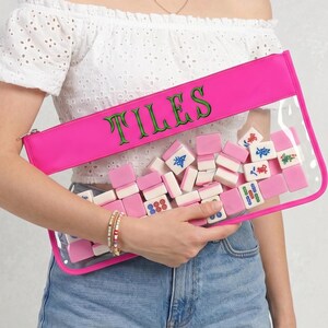 May include: A clear pouch with a vibrant pink trim and zip, containing pink and white mahjong tiles. The word "TILES" is embroidered in green on the pink top. The pouch is held by a person wearing a white off-the-shoulder top and blue denim jeans.