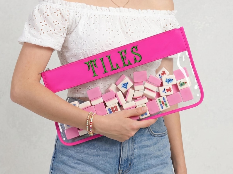 May include: A clear pouch with a vibrant pink trim and zip, containing pink and white mahjong tiles. The word "TILES" is embroidered in green on the pink top. The pouch is held by a person wearing a white off-the-shoulder top and blue denim jeans.