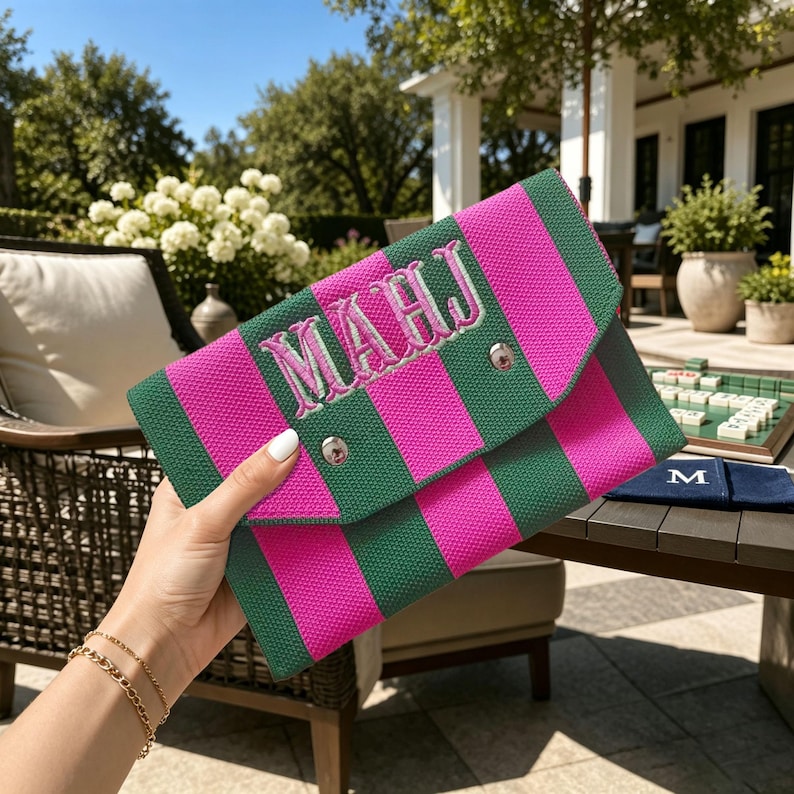 May include: A rectangular clutch bag featuring vertical stripes in vibrant hot pink and dark forest green. The front flap is secured with a silver metal snap button and displays the embroidered monogram letters MAHJ in a light pink thread.