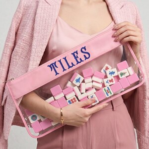 May include: A pink pouch with the word "TILES" embroidered in blue. The clear front of the pouch displays a collection of pink and white Mahjong tiles. The pouch is held by a person wearing a pink outfit.