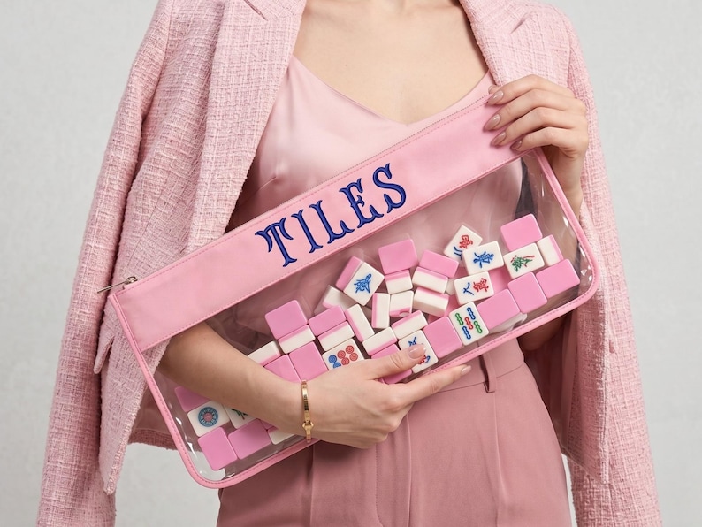 May include: A pink pouch with the word "TILES" embroidered in blue. The clear front of the pouch displays a collection of pink and white Mahjong tiles. The pouch is held by a person wearing a pink outfit.