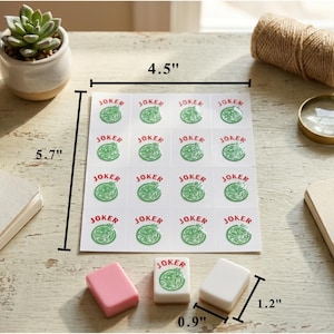 May include: A collection of game pieces and accessories on a wooden surface. A 4.5" x 5.7" sheet of stickers with "JOKER" text and a green graphic is present. Three game pieces are also visible, along with a magnifying glass and a small succulent plant.