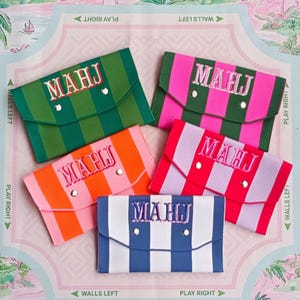 May include: Five rectangular envelope-style pouches with striped patterns in green, pink, orange, red, and blue. Each pouch features a flap with two silver-toned snap buttons and the embroidered text MAHJ in a contrasting thread color on the front.