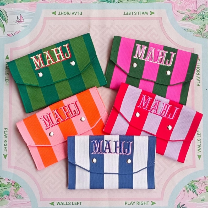 May include: Five rectangular envelope-style pouches with striped patterns in green, pink, orange, red, and blue. Each pouch features a flap with two silver-toned snap buttons and the embroidered text MAHJ in a contrasting thread color on the front.