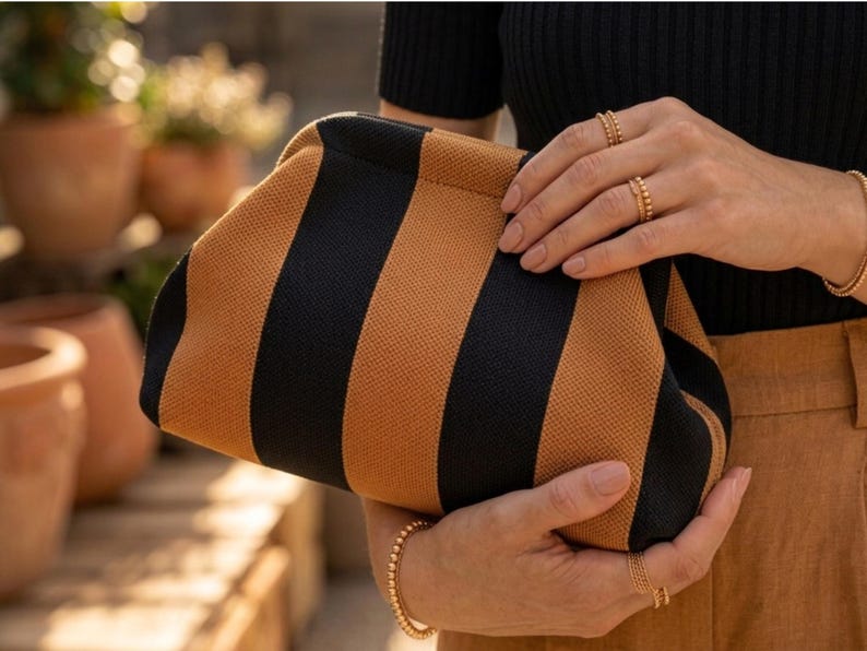 May include: A structured clutch bag featuring a bold, vertical striped pattern in alternating black and tan colors. The bag has a soft, woven fabric texture and a curved, frame-style top closure, providing a clean and simple aesthetic design.