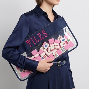 May include: A navy blue pouch with a clear front, filled with pink and white mahjong tiles. The word "TILES" is embroidered in pink on the navy blue border. The pouch is rectangular and held by a person wearing a navy blue outfit.