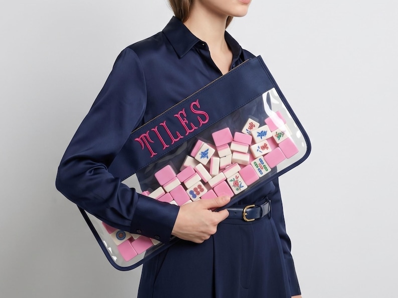 May include: A navy blue pouch with a clear front, filled with pink and white mahjong tiles. The word "TILES" is embroidered in pink on the navy blue border. The pouch is rectangular and held by a person wearing a navy blue outfit.