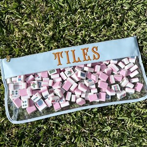May include: A light blue pouch filled with pink and white tiles. The word "TILES" is embroidered in gold thread across the top. The pouch has a clear front, and a gold zip. The background is green grass.
