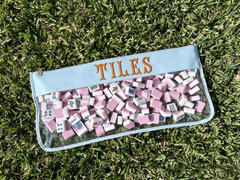 May include: A light blue pouch filled with pink and white tiles. The word "TILES" is embroidered in gold thread across the top. The pouch has a clear front, and a gold zip. The background is green grass.