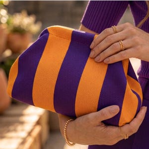 May include: A clutch bag featuring a bold, vertical striped pattern in alternating shades of deep purple and vibrant orange. The bag has a soft, fabric-like texture and a rounded, structured frame that is held securely in place by two hands.