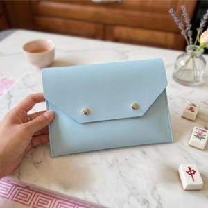 May include: A pale blue clutch bag with a flap and two gold-coloured stud fasteners. The rectangular bag rests on a marble surface. Mahjong tiles, a small vase with flowers, and a teacup are also visible.