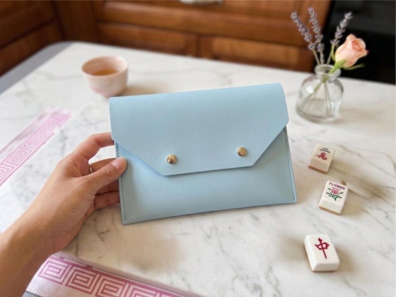May include: A pale blue clutch bag with a flap and two gold-coloured stud fasteners. The rectangular bag rests on a marble surface. Mahjong tiles, a small vase with flowers, and a teacup are also visible.