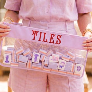 May include: A clear pouch with the word "TILES" embroidered in red. The pouch is filled with small, rectangular tiles in shades of lavender, white, and orange. The tiles feature various illustrations. The pouch is held by a person wearing a pink outfit.